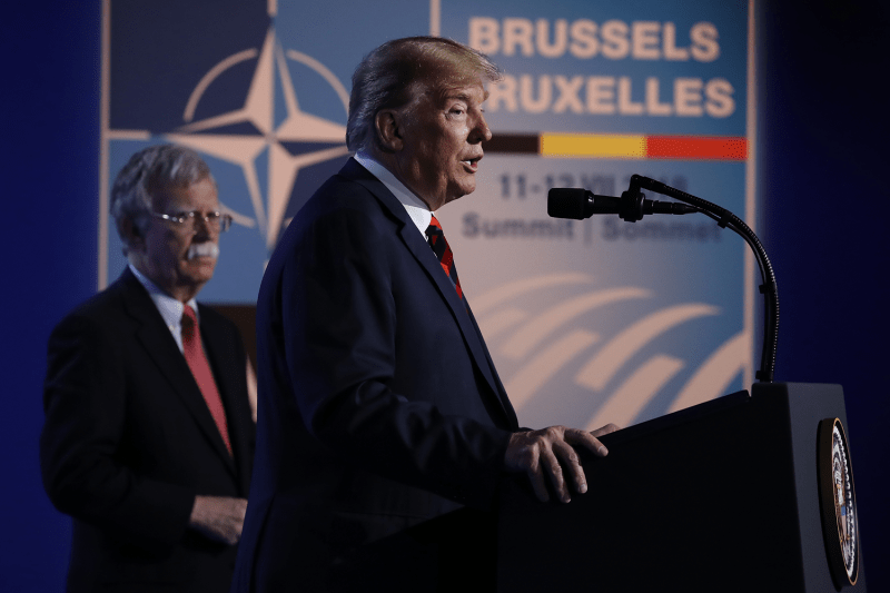 U.S. President Donald Trump is seen in profile as he stands at a podium and speaks into the microphone at NATO summit. His national security advisor stands slightly behind him, watching the crowd. Both men wear dark suits and ties.