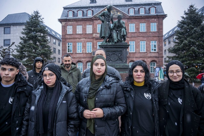 Women wearing black, some with headscarves, stand looking forward in front of a statue in Germany. Behind them is a crowd of other men and women.