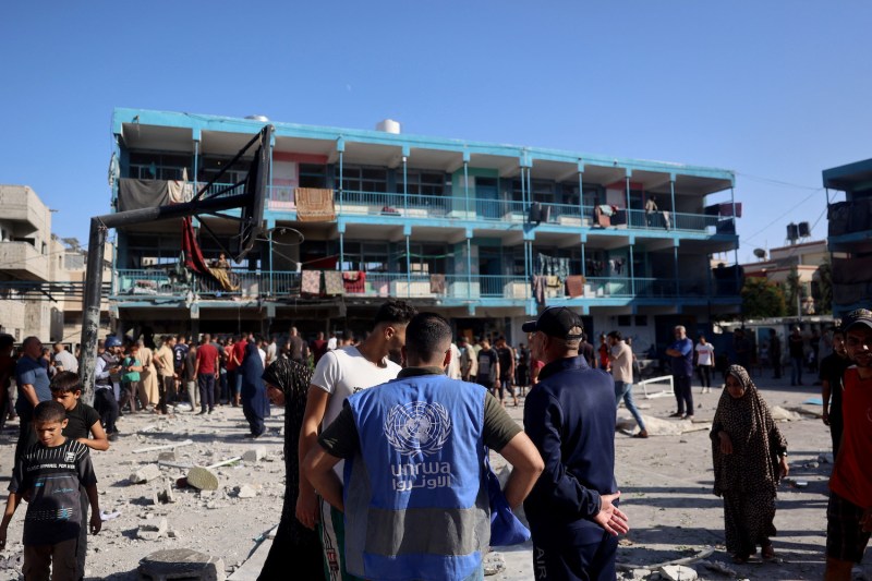 Dozens of people mill about the remains of a rubble-covered basketball court in front of a three-story building with a blue awning. One of the closest people, facing away from the camera, wears a blue vest with the U.N. logo and "UNRWA" printed on the back.