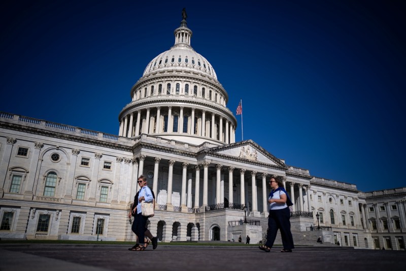 The white marble dome of the U.S. Capitol building looms over a deep blue sky. Several people can be seen in the foreground as they walk across the plaza in front of the building.