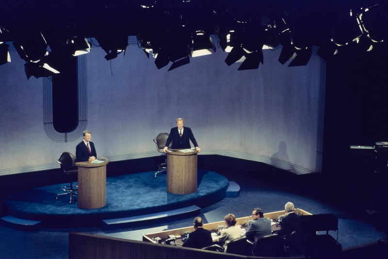 Stage lights are seen above a stage with two men behind lecturns. In front of them are four people behind a dais looking toward the stage.