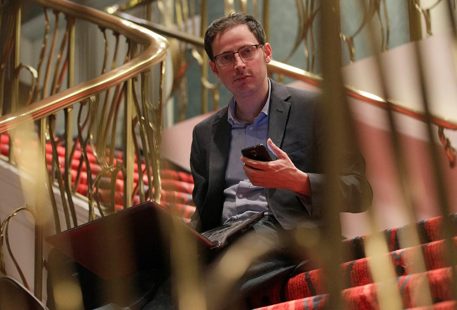 Nate Silver in a suit jacket and button-up shirt sits on stairs surrounded by gold railing holding a cell phone.