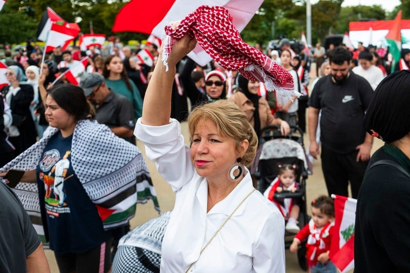 A woman in a white button up shirt waves a red and white keffiyah above her head. Behind her are a crowd of people holding Lebanese and Palestinian flags. Just over her shoulder is a stroller and two young children framed by flags.