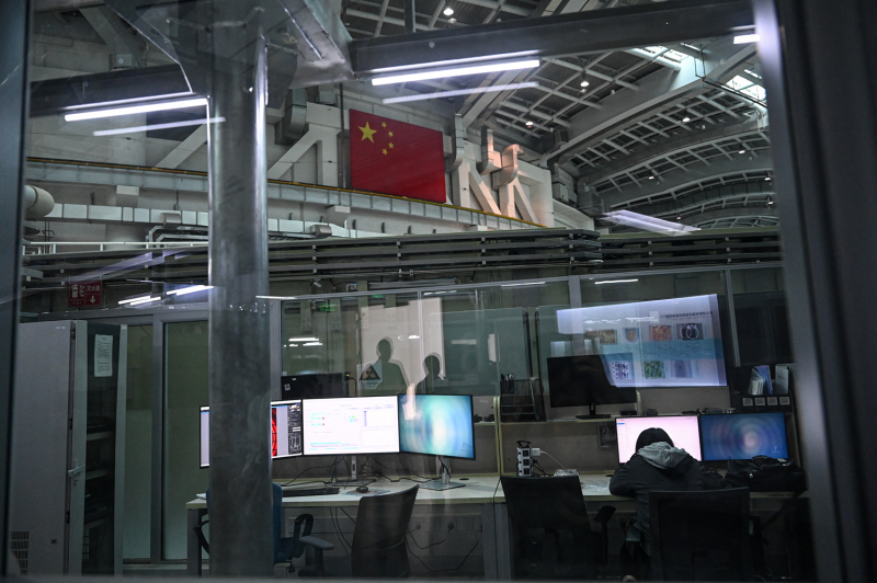 A worker sitting at a desk with several computer monitors is seen from behind and across a room in a facility in Shanghai. Other monitors hang above nearby work desks, and a walkway or scaffolding stretches overhead. A Chinese flag hangs from high up on a nearby wall.