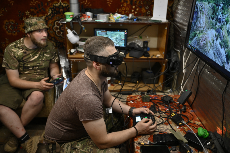 Two Ukrainian servicemen dressed casually in T-shirts sit in a small room crowded with electronic cables and other devices. The man closer to the camera is seen in profile with a headset on and a controller in his hands as he operates a drone using a monitor screen to the right. The other man sits farther back, holding a controller as well as he supports his comrade.