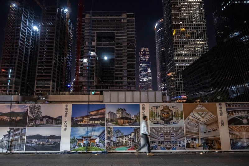 A pedestrian walks on a street in Beijing at night beneath a dark sky, passing by a short wall covered by advertisements while skyscrapers loom in the background. The ads are covered with photos of various buildings and architectural details.