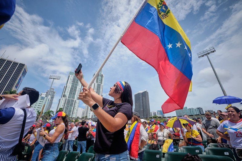 A woman holds up her phone to take a photo as she holds a large Venezuelan flag above her head. Behind her is a large crowd and the skyline of Miami.