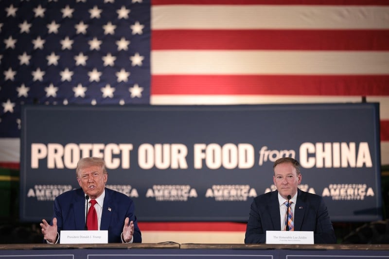 Donald Trump gestures as he sits at a table behind a microphone. At right is another man seated. Behind them is the U.S. flag and a sign that says "Protect Our Food from China."