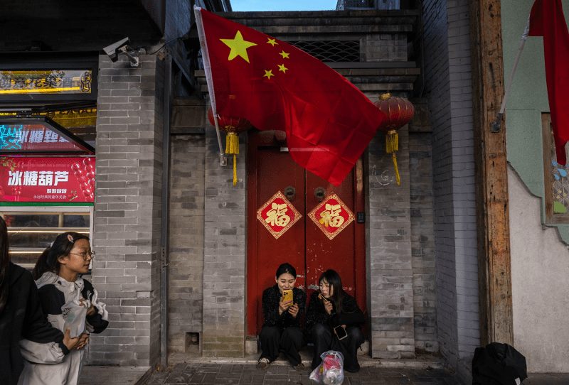 Two people sit on the steps in front of a red door in Beijing. Above them is the Chinese flag.