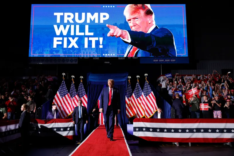 Donald Trump on a stage at a campaign rally. Behind him are multiple American flags and an image of him with the words "Trump will fix it."