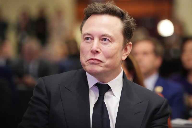 Elon Musk is seen from the chest upward as he sits among a crowd of people at a meeting. Musk wears a black suit and tie and an expression that makes it look like he's somehow smiling and frowning at the same time.