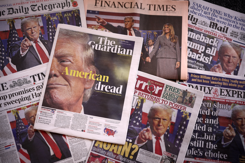 British newspapers including the Guardian, the Telegraph, the Financial Times, and the Mirror are scattered on a tabletop overlapping each other. Each has a photo of U.S. President-elect Donald Trump on the page; accompanying headlines include "American dread" and "Again?"