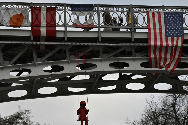 People walk on a bridge past an American flag and above an art installation by French street artist James Colomina called the "Swing" which depicts a little girl wearing an oversized military helmet as sits on a playground swing hanging from the bridge.