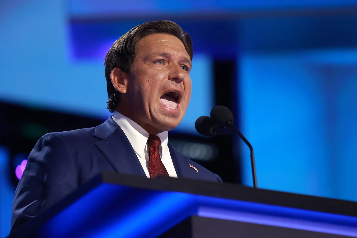 Ron DeSantis opens his mouth wide as he talks behind a microphone and lectern in front of a blue screen.