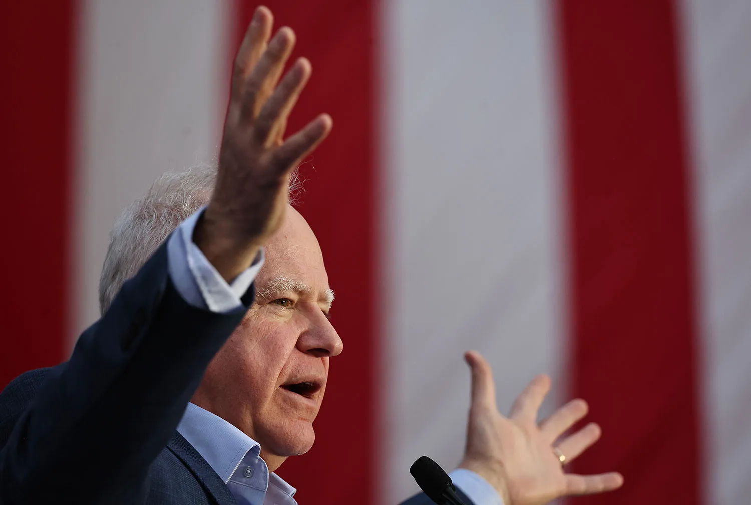 Tim Walz holds up both hands as he talks into a microphone. A U.S. flag is seen behind him.