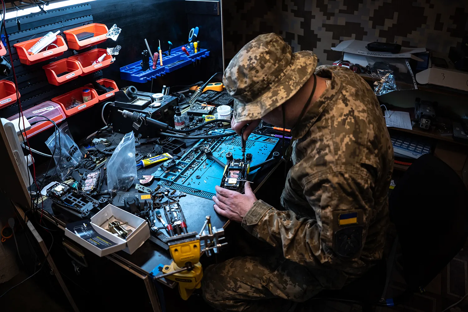 A man in military camouflage sits at a table working on electronic components with a tool.