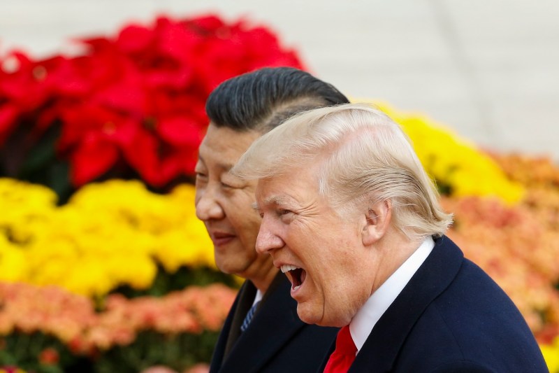 U.S. President Donald Trump reacts with his mouth wide open as he takes part in a welcoming ceremony with Chinese President Xi Jinping, who stands beside him. Piles of yellow, orange, and red flowers are visible behind the men.