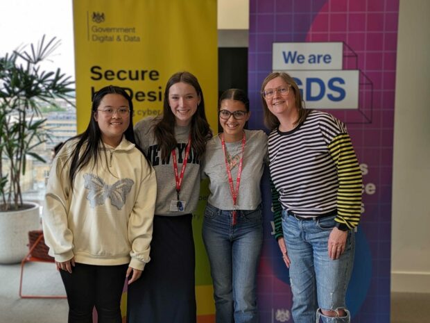 The image features three young women from Next Tech Girls, alongside a member of the GDS team, Dr Sarah Kirby-Ginns. They're standing in front of two pop-up banners, smiling.