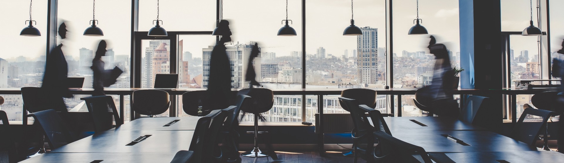 widescreen view of a busy office in motion. In the foreground, a series of empty desks. In the midground, people in business attire are walking by and appear blurry. In the background, a city skyline appears clearly though the sky looks white and cloudy.