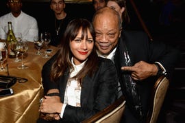 rashida jones and quincy jones smile for a photo while seated at a table together, he points at her with one hand as they holds hands, both wear black and white outfits