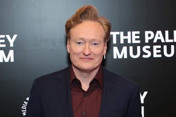 conan obrien smiles at the camera, he wears a navy suit jacket and red collared shirt