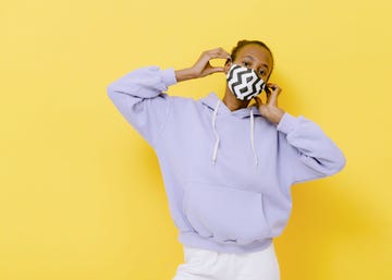 a young girl stands in front of a yellow background and wears a protective mask during the coronavirus she wears lavender hoodie and white jeans and her elbows are wide apart