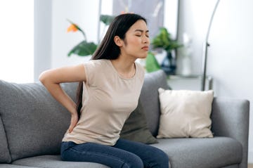 upset frustrated asian young woman suffering from backache, unhappy japanese brunette sitting on a sofa in living room, feeling discomfort because of pain in back, need treatment and rest