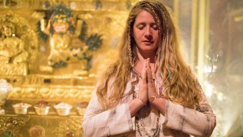 guru jagat, aka katie briggs, holds her hands together at her chest and closes her eyes, she wears a light colored long sleeve shirt and necklaces, a golden ornate setting is in the background