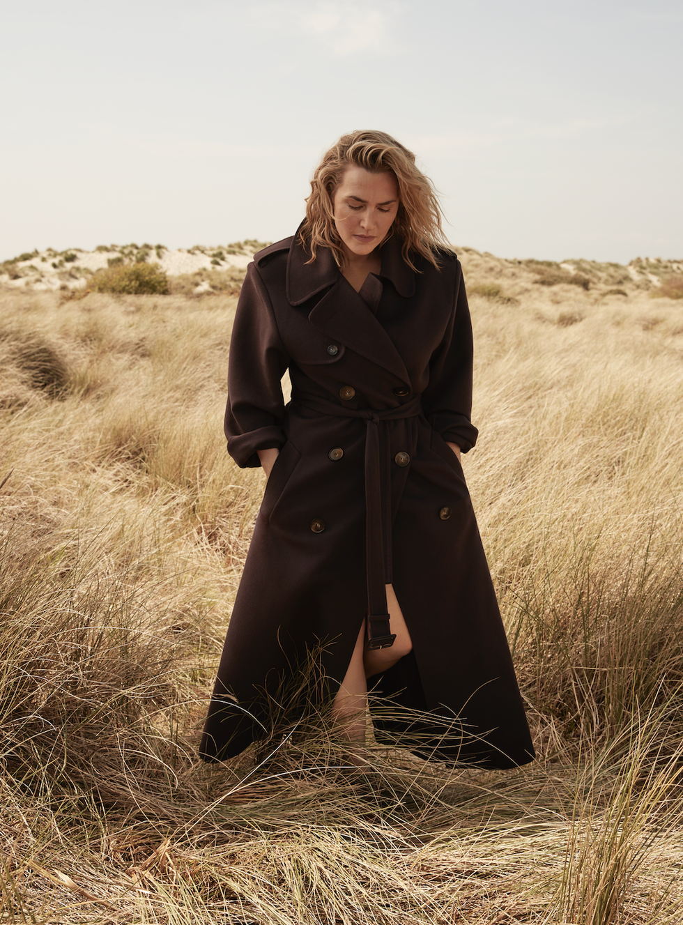 kate winslet stands in a grassy field in a coat