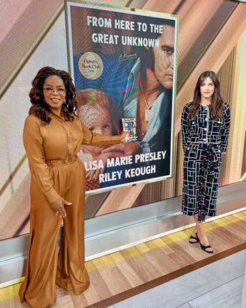 the scene features two women standing next to a prominent book cover displayed in the background the book, highlighted as part of oprah's book club, is a memoir by lisa marie presley and riley keough the woman on the left is wearing a brown outfit with a belt, holding a copy of the book, while the woman on the right is dressed in a patterned black and white suit