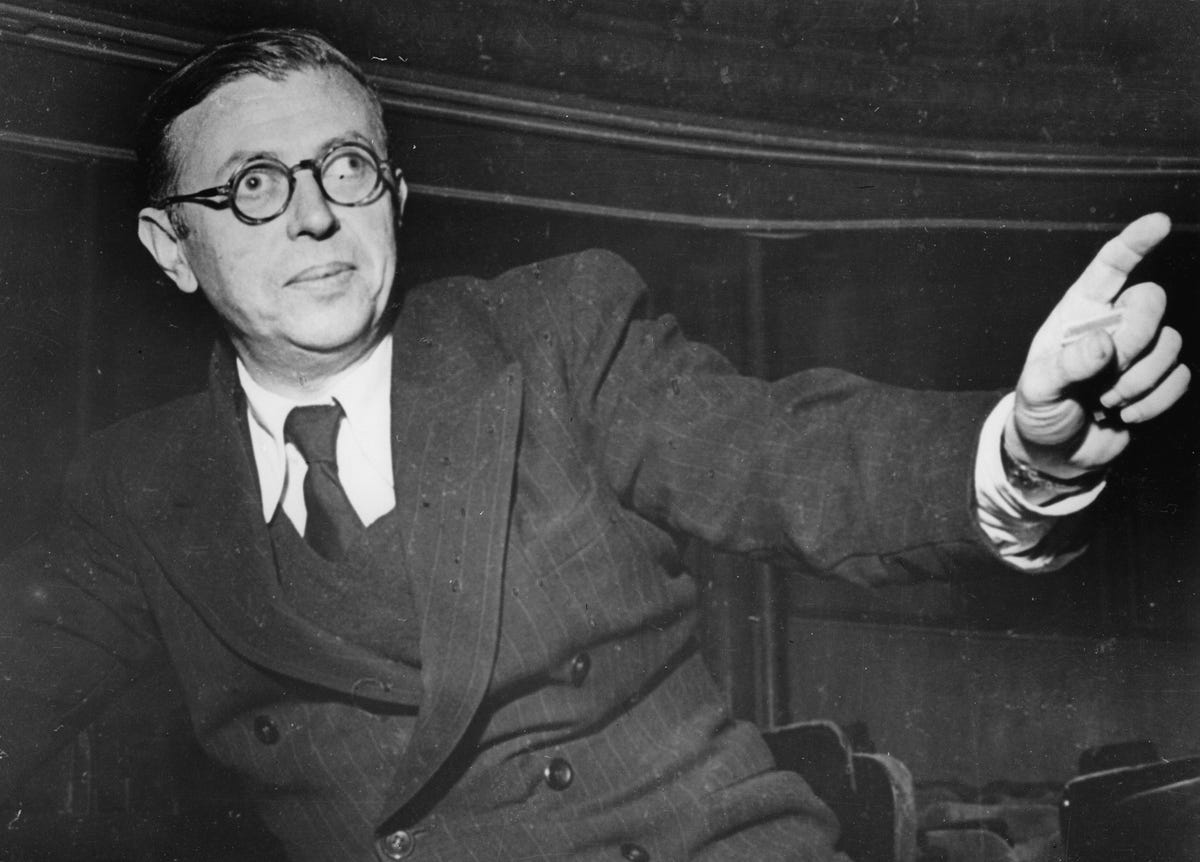 jean paul sartre sits in a three piece suit and tie and points one finger out to the right