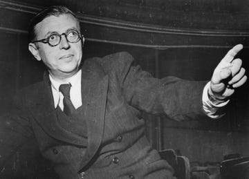 jean paul sartre sits in a three piece suit and tie and points one finger out to the right