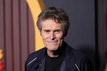 willem dafoe smiles at the camera, he wears a dark jacket over a black shirt