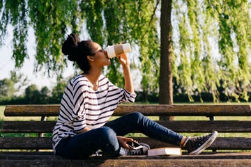 young woman sitting on park bench drinking coffee to go