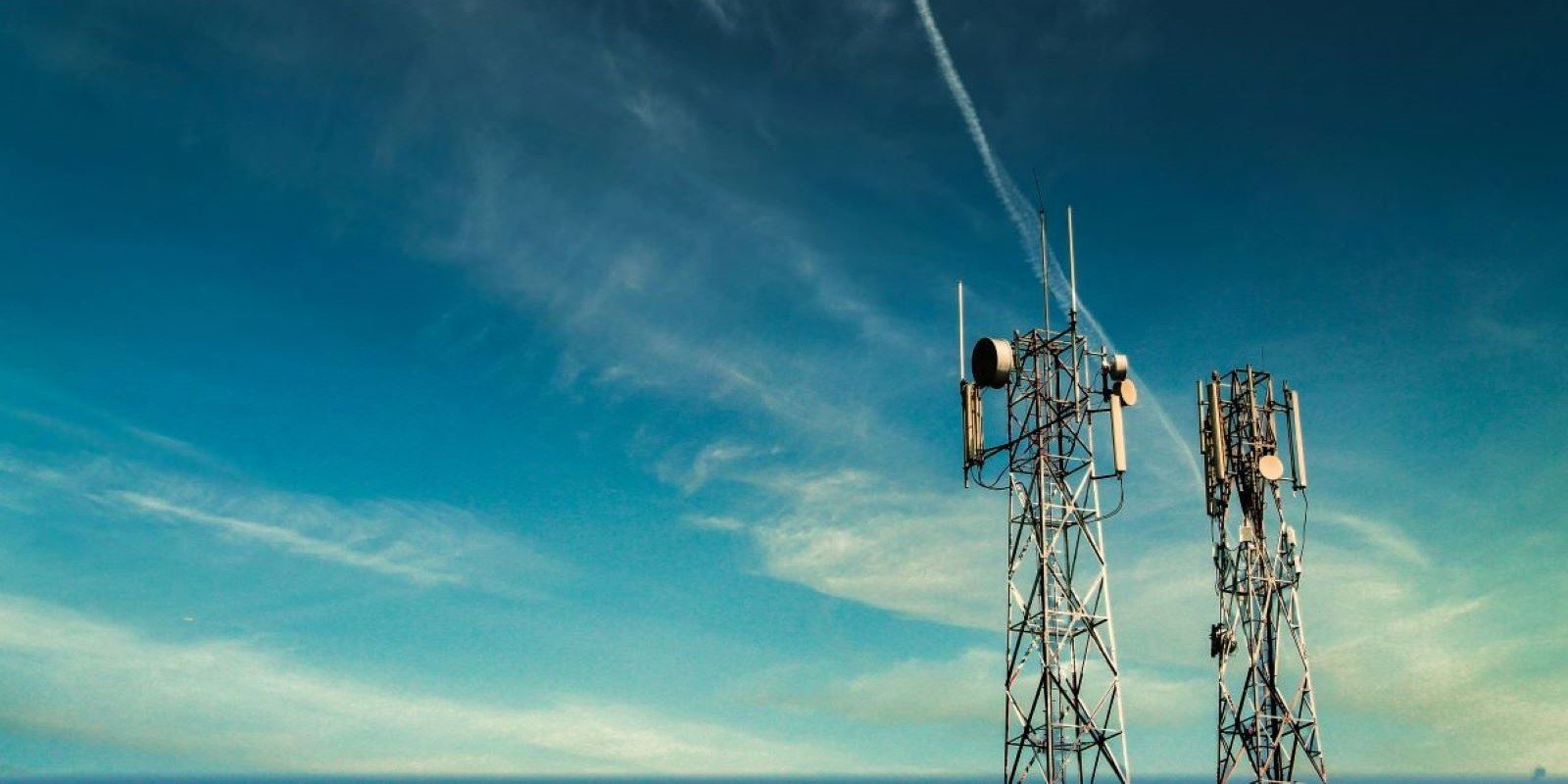 Two cellular network towers stand against a bright blue sky with wispy clouds, showcasing modern telecommunication infrastructure.