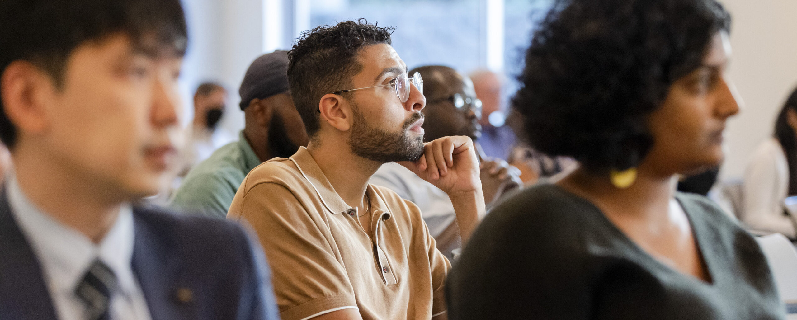 A young man sits and looks off-camera, there are two other people closer to the camera that are not in focus.