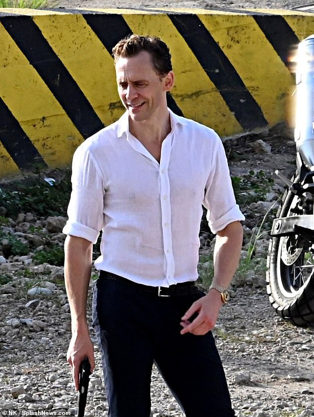 Tom Hiddleston smiles with a gun in hand as he appears on set of The Night Manager