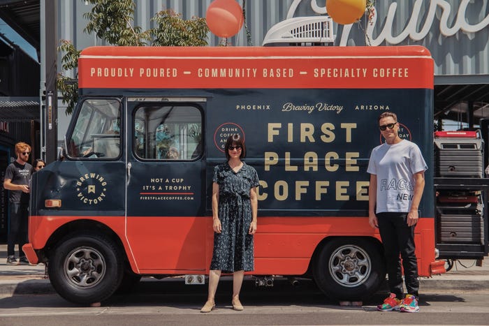 Photo of two people standing in front of a red and black truck that reads "First Place Coffee" in large tan lettering. There are two balloons above the truck.