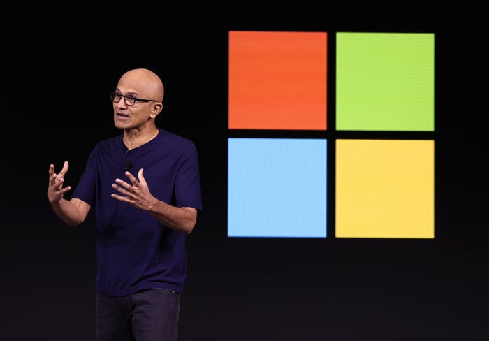 Satya Nadella standing in front of a Microsoft logo.