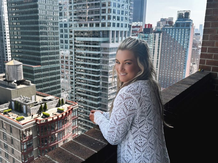 Sarah Michelle Sherman wearing a white crocheted sweater and standing on a balcony in a city.