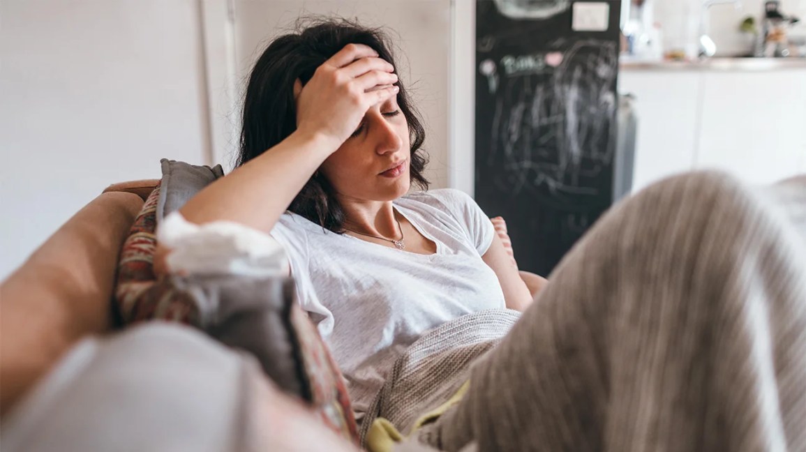 A woman sits on a couch, holding her hand to her head. 