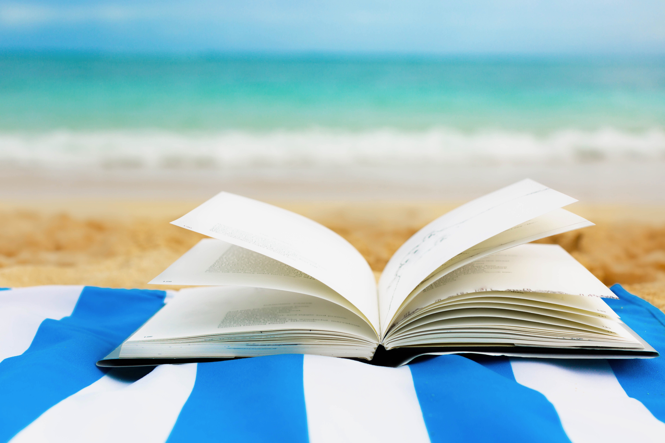 A book opened lying on a blue towel on the sand on the beach.