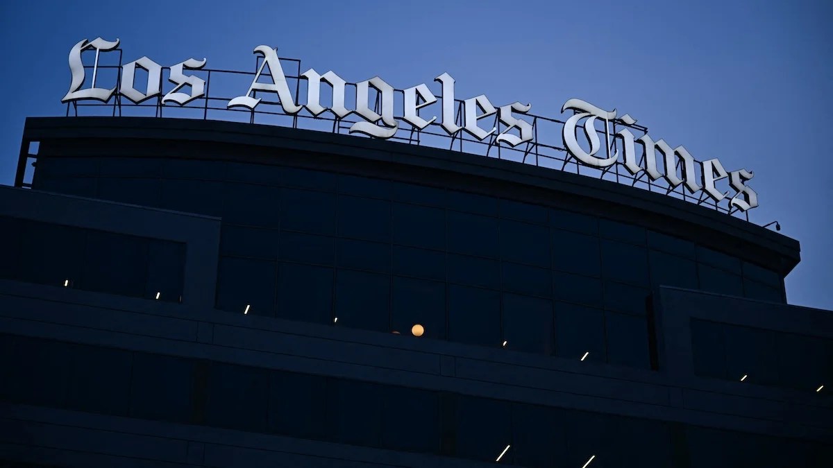 The Los Angeles Times' white neon sign is illuminated in the light of dusk, a dim blue sky behind it, atop the Times' El Segundo headquarters building below.