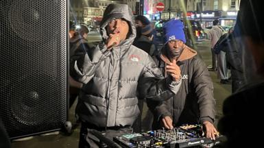 A man raps behind a set of DJ decks set up on a street at night. He is wearing a hooded puffer jacket. Beside him a man operates the decks, he is wearing a dark hooded puffer jacket with a blue hat with an AG logo. To the left of them is a large speaker