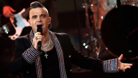 Robbie Williams wearing a bejewelled black jacket and a black top, singing into a microphone with a band behind him 