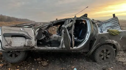 A burnt out pick-up truck in a field full of mud and rubble. One of the doors is inside out and the inside of the vehicle is destroyed