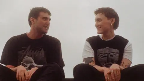 Fred Again and SOAK, both wearing black t-shirts and trousers, sitting on a ledge with grey sky in background