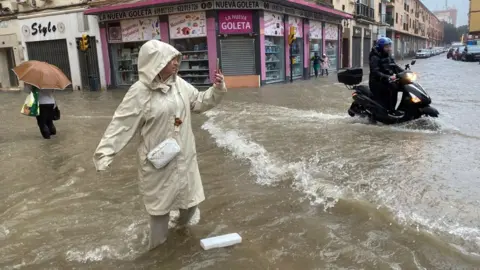 A woman, wearing an overcoat and knee deep in water, takes a photo of a flooded street in Malaga.  Behind her a man on a motorcycle ploughs through the water.