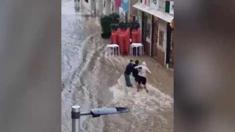 Three people help each other walk through water flooding down a street.