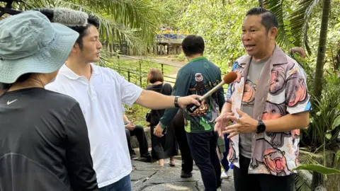 Khao Kheow Open Zoo/X Zoo director Narongwit Chodchoi, wearing a hippo patterned shirt, being interviewed by a Japanese television crew.